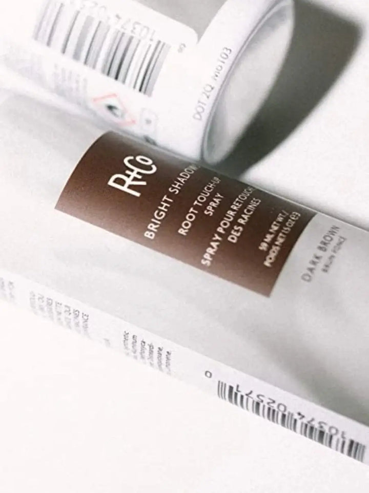 A close-up shot focuses on the sleek label details of an R+Co BRIGHT SHADOWS Root Touch Up Spray - Dark Brown bottle laying on a clean white surface | Twentyseven Toronto