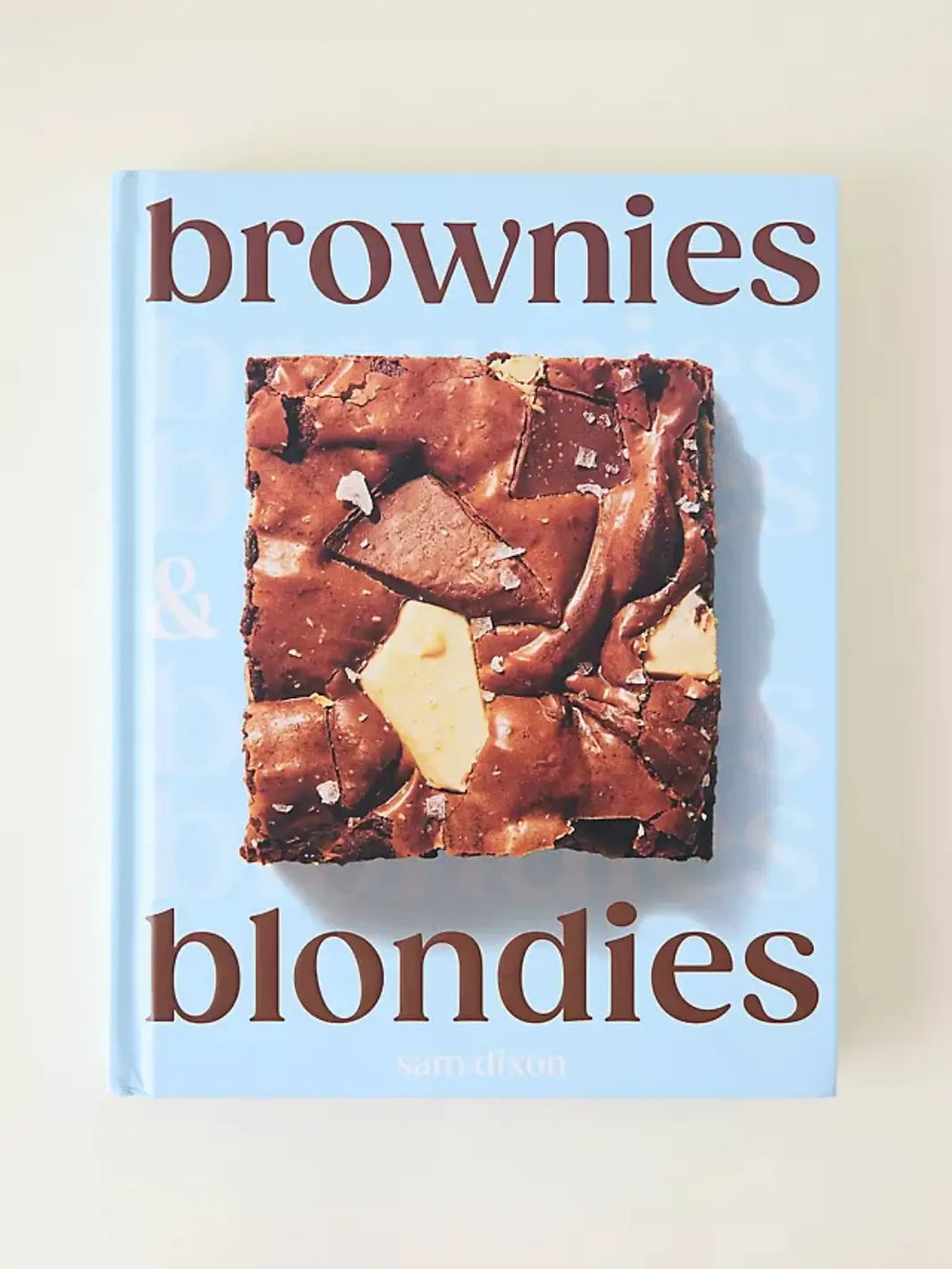 Front cover of the cookbook "Brownies & Blondies" by Sam Dixon featuring light blue with large brown text and close up photograph of a thick, square brownie with large chocolate chunks and a sprinkle of sea salt | Twentyseven Toronto