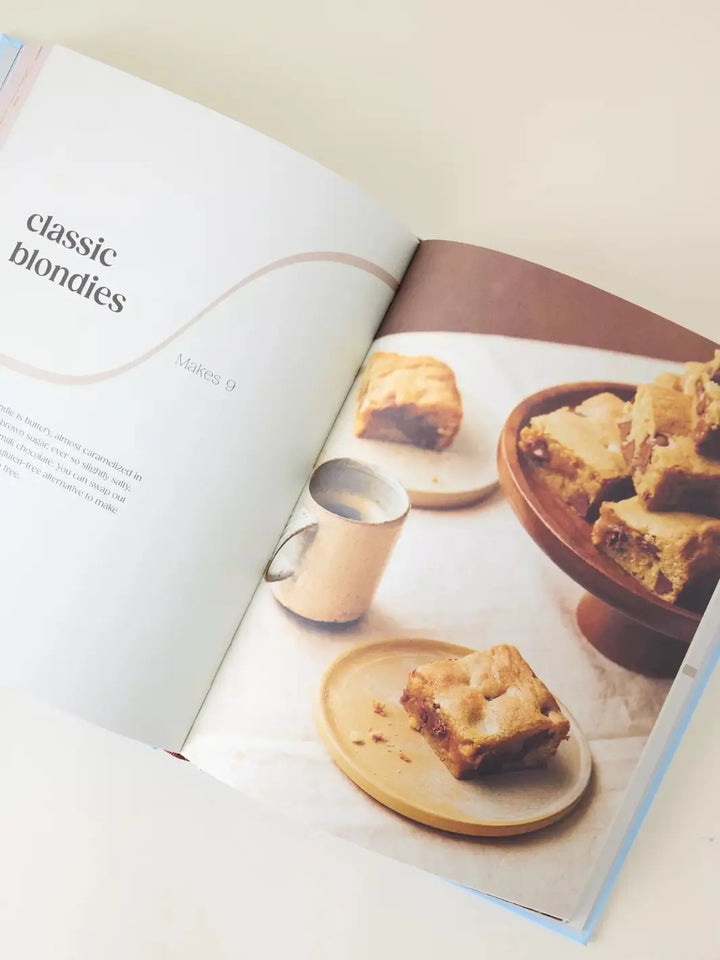 An open cookbook spread titled classic blondies from Brownies and Blondies book by Sam Dixon. The right page features a lifestyle photograph of several golden blondies with chocolate chips served on small wooden plates and a pedestal stand, accompanied by a small mug of coffee | Twentyseven Toronto