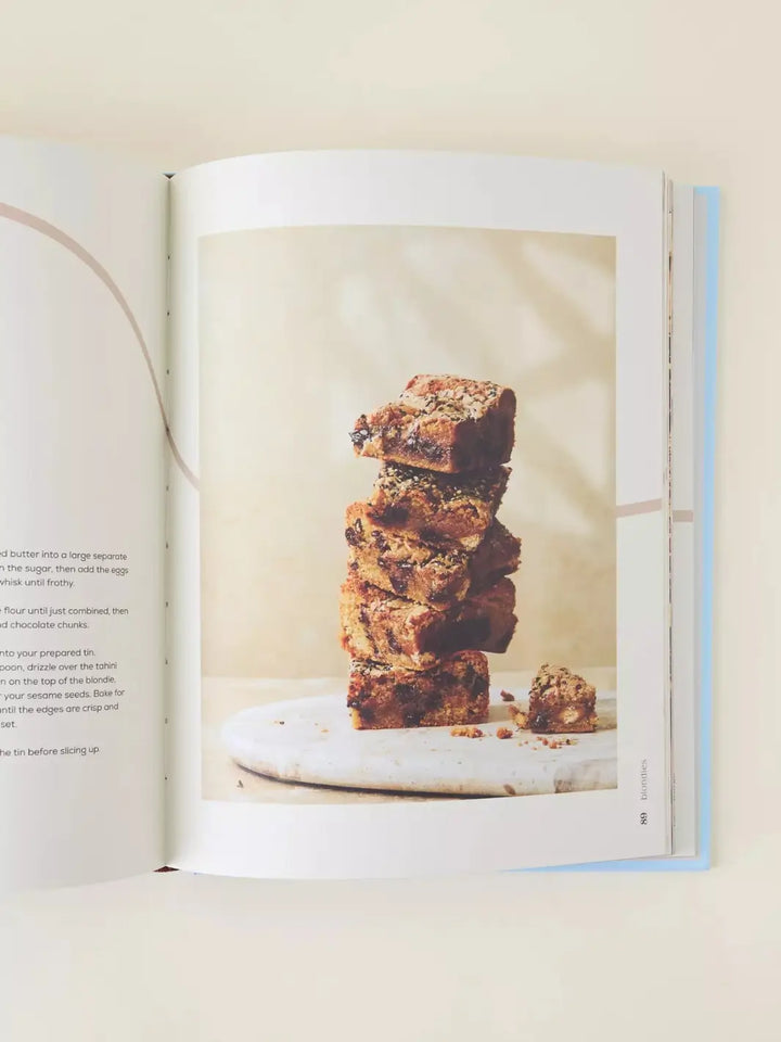 An open cookbook page showing a vertical stack of five blondies on a marble surface, highlighting the gooey texture and chocolate inclusions | Twentyseven Toronto
