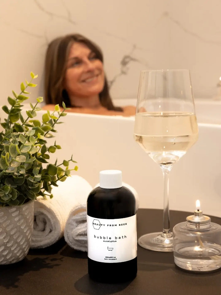 Woman in a bathtub with a bottle of bubble bath in Eucalyptus from Beauty from Bees, a glass of wine, and a candle | Twentyseven Toronto
