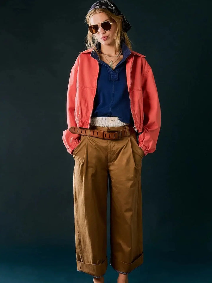 A full-length front view shows a model showcasing the relaxed, pleated fit of the Free People Buckle Back Trousers - Tobacco Combo paired with a bright coral jacket | Twentyseven Toronto