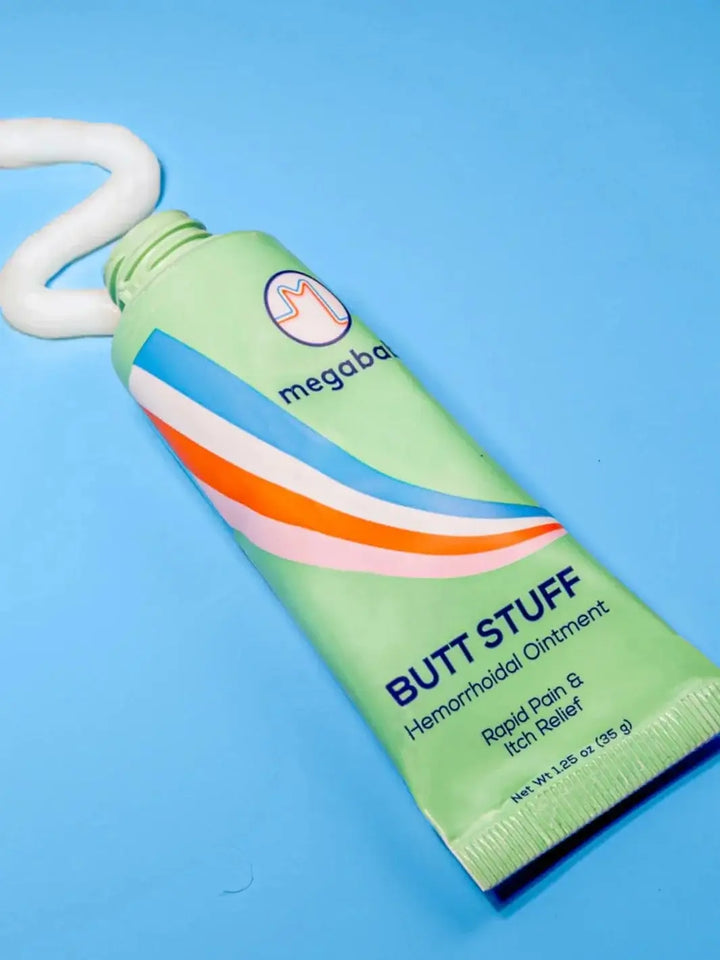 A light green squeeze tube is shown against a bright blue background with a dollop of white ointment squeezed out from the top, highlighting the texture of the Megababe Butt Stuff Hemorrhoid Cream | Twentyseven Toronto