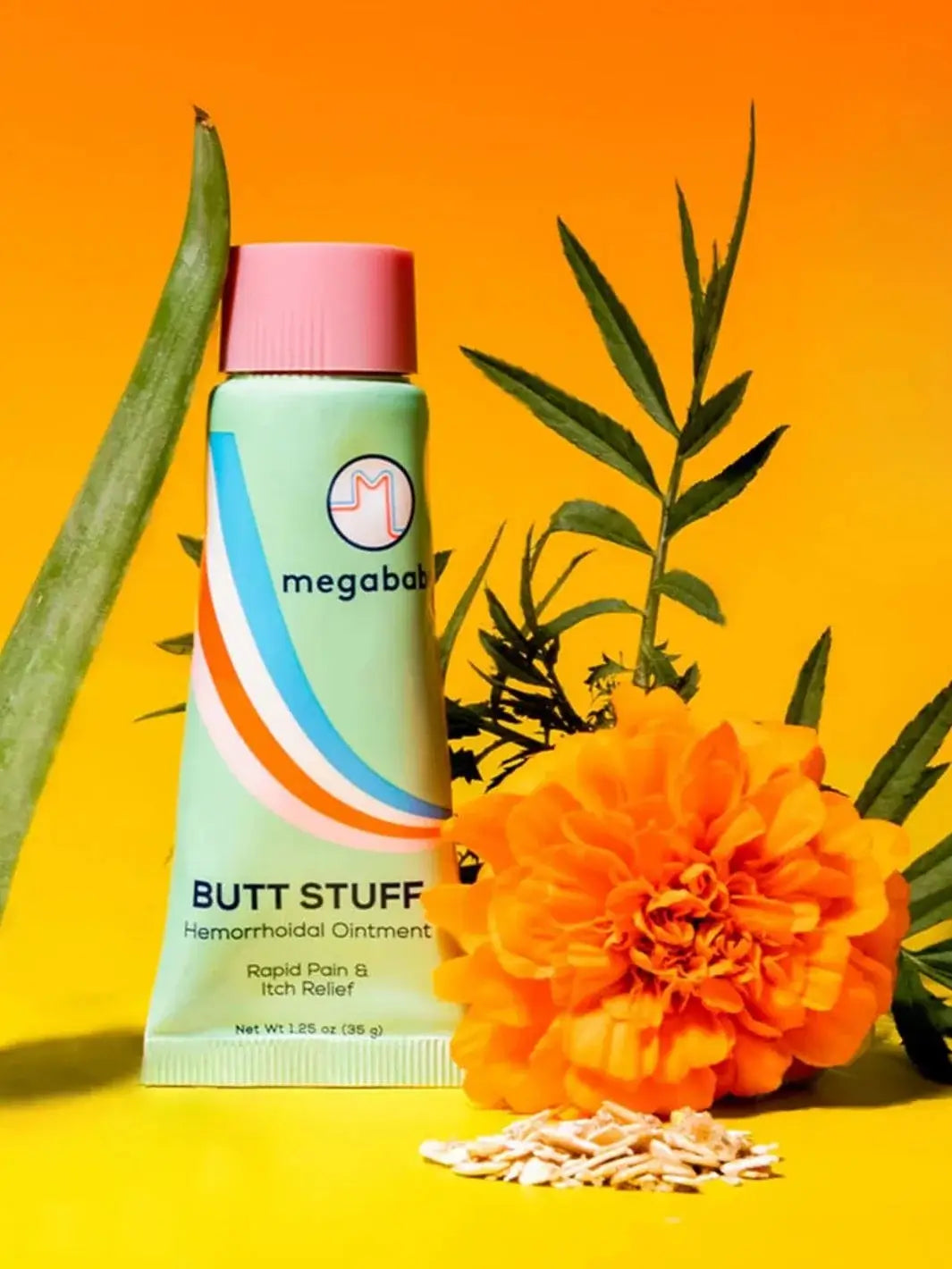A mint green ointment tube stands vertically against a solid yellow backdrop flanked by an aloe leaf and an orange marigold flower, emphasizing the soothing ingredients in the Megababe Butt Stuff Hemorrhoid Cream | Twentyseven Toronto