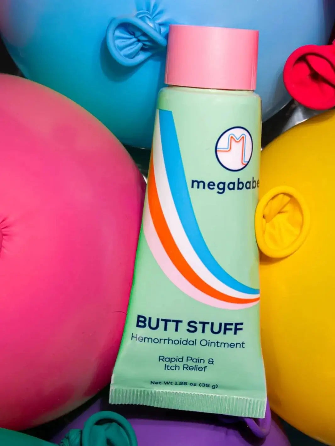 A light green skincare tube featuring colorful stripes is nestled closely among vibrant pink, yellow, and blue balloons to showcase the playful packaging style of the Megababe Butt Stuff Hemorrhoid Cream. | Twentyseven Toronto
