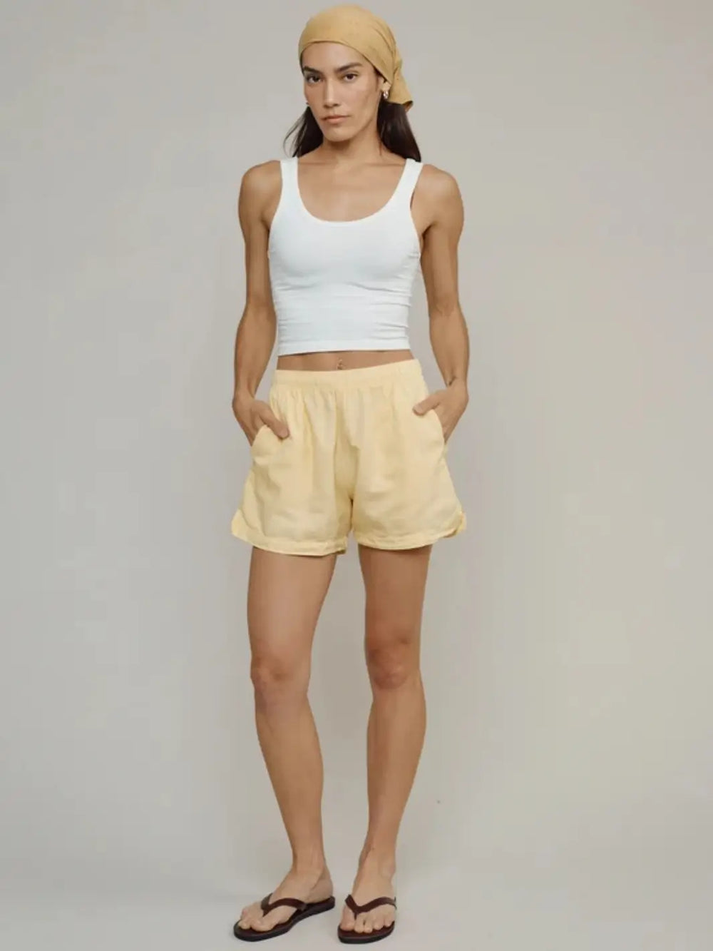 Full-length front view of a model wearing butter yellow Jungmaven Cabana Shorts paired with a white tank top and a yellow headscarf | Twentyseven