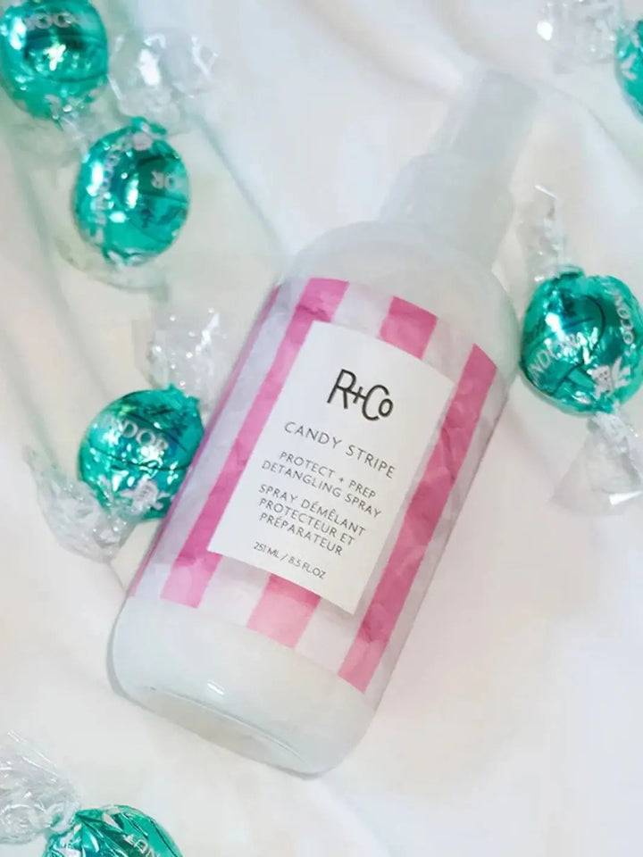 A spray bottle of R+Co CANDY STRIPE Protect + Prep Detangling Spray lies surrounded by several wrapped teal chocolate candies | Twentyseven Toronto