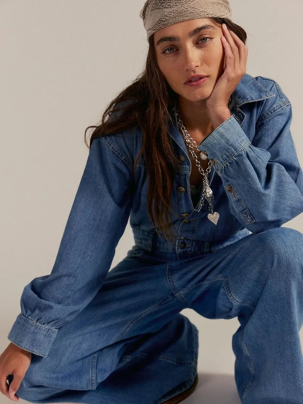 A model crouches down to show the relaxed collar, buttoned front, and long sleeves of the medium wash denim Free People Carpe Diem One-Piece in New Day | Twentyseven Toronto