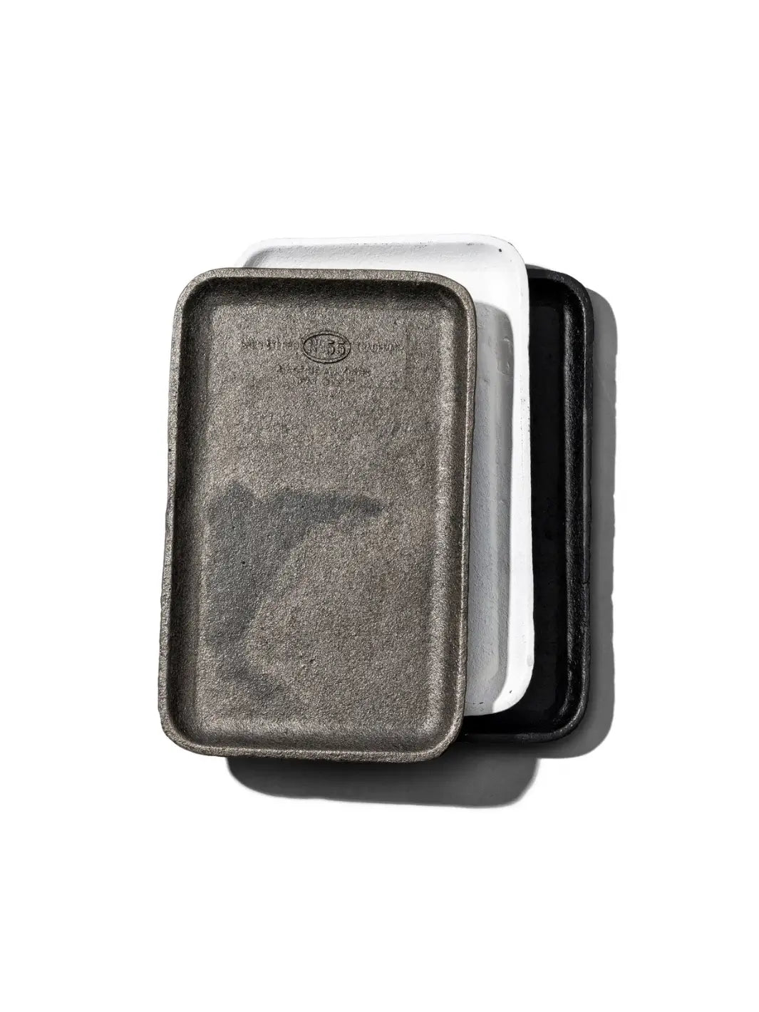 A top-down view shows three rectangular dishes stacked together, featuring the Cast Iron Tray collection from Puebco at the very bottom beneath white and natural grey variations | Twentyseven Toronto