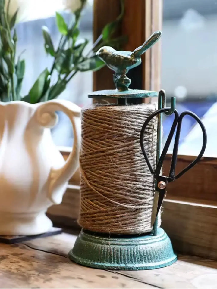 A rustic Cast Iron Twine Dispenser with Bird Accent from Laura Hooper Design House features a decorative bird perched on top and a spool of twine held within its vintage-style frame | Twentyseven Toronto