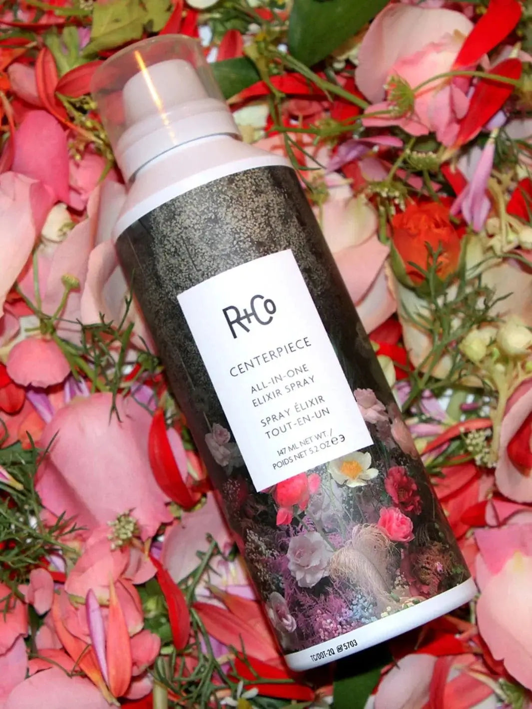 A spray bottle of R+Co CENTERPIECE All-In-One Elixir Spray rests flat against a vibrant bed of colorful flower petals | Twentyseven Toronto