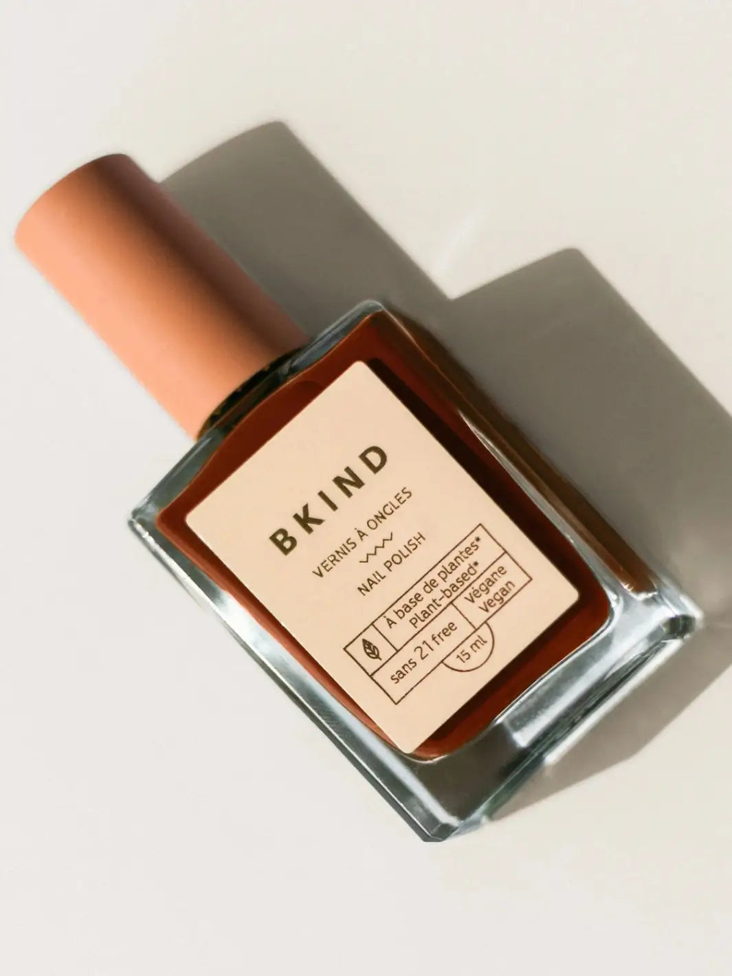 A square glass bottle of BKIND Nail Polish in Chai with a beige cap rests diagonally against a clean, off-white backgroun | Twentyseven Toronto