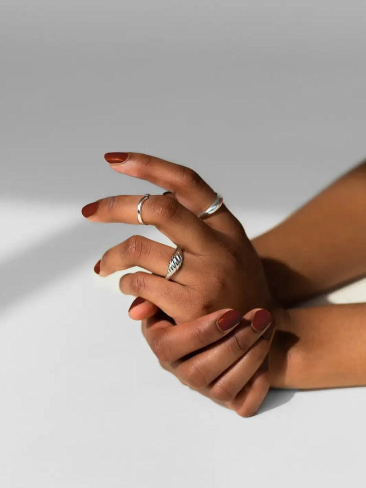 Hands adorned with silver rings are crossed, showcasing a glossy, deep terracotta-brown manicure featuring BKIND Nail Polish in Chai | Twentyseven Toronto