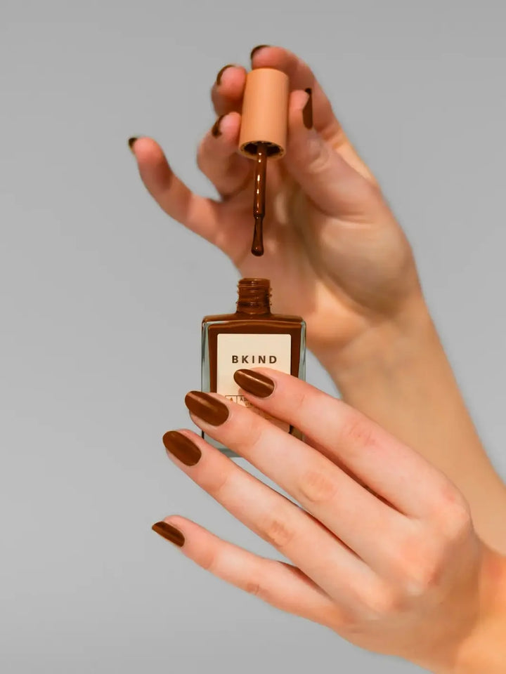 A pair of hands holds open a bottle of BKIND Nail Polish in Chai, with the brush lifting out a droplet of the rich, warm brown liquid over a matching manicure | Twentyseven Toronto