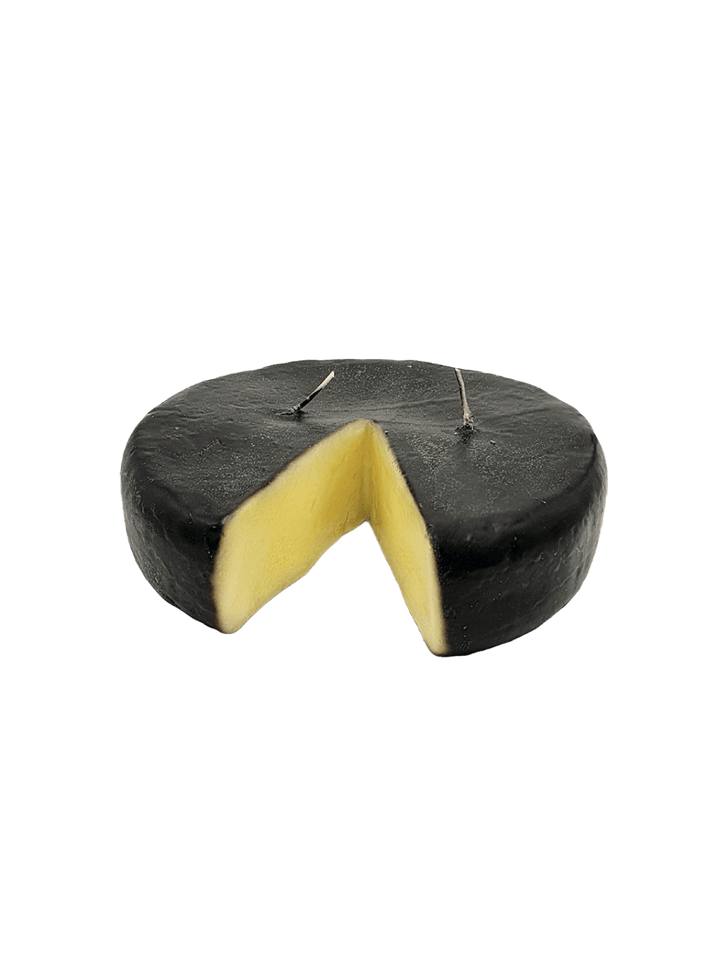 Black rind cheese wheel shaped candle from Cereria Introna with a wedge cut out on a transparent background | Twentyseven Toronto
