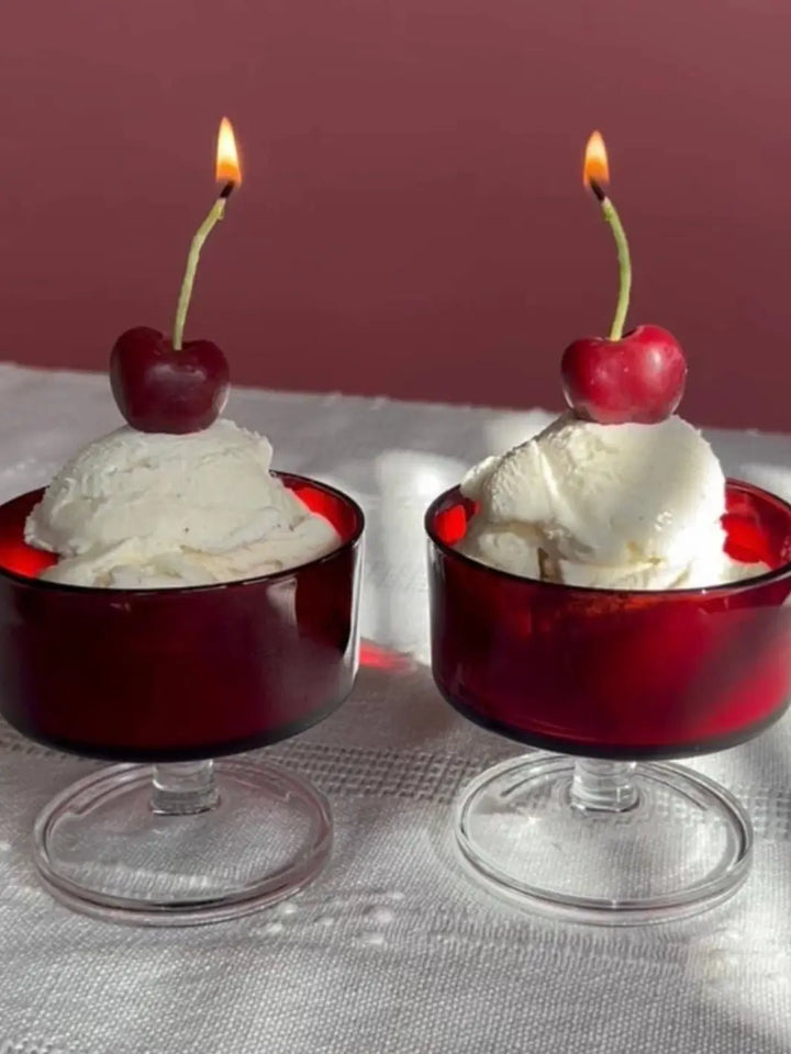 Two red glass bowls with vanilla ice cream, and lit cherry-shaped candles from Happy Organics on a table and red background | Twentyseven Toronto