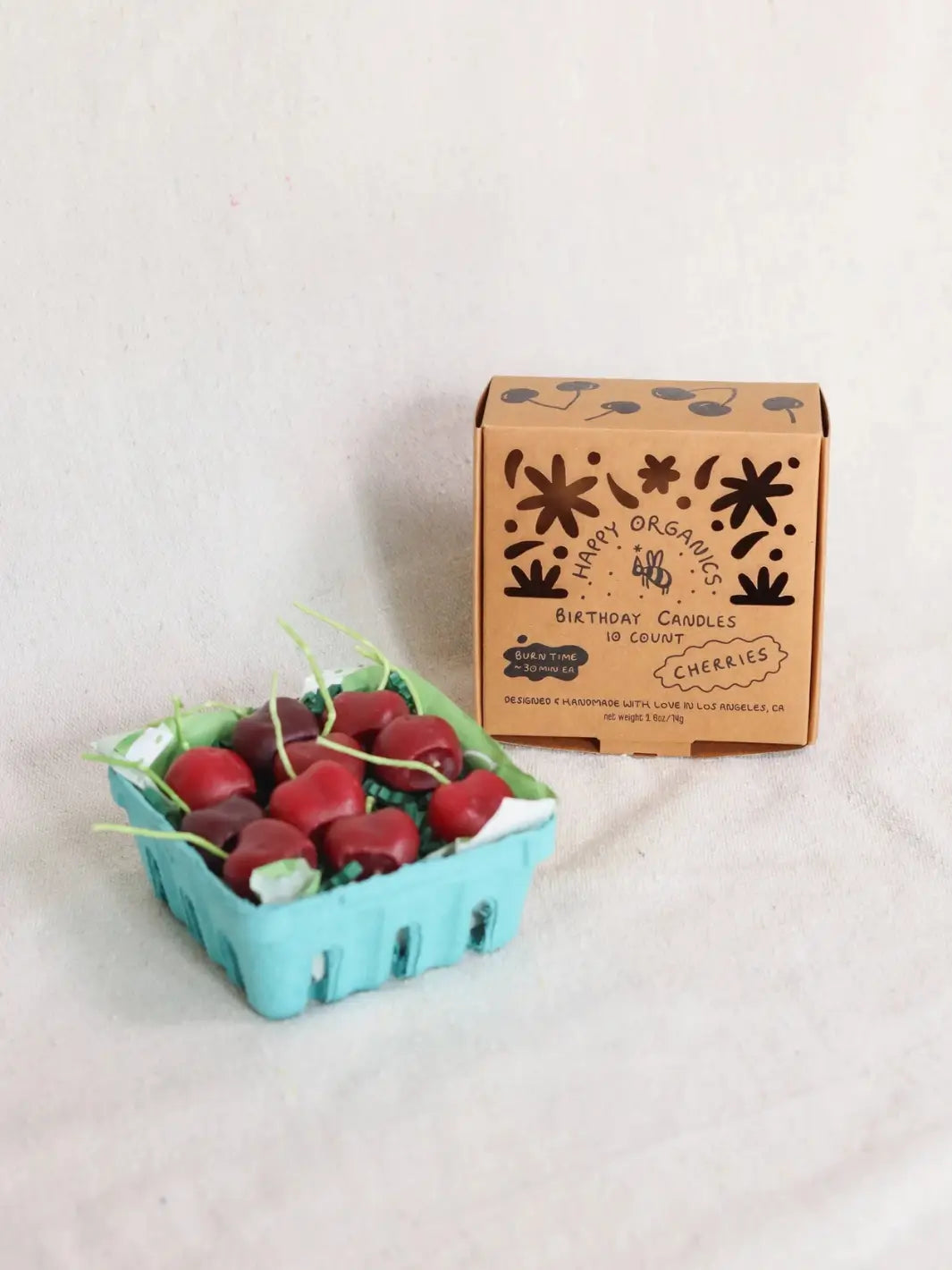 Carton of red birthday candles shaped like cherries next to a box labeled 'Happy Organics' on a light background | Twentyseven Toronto