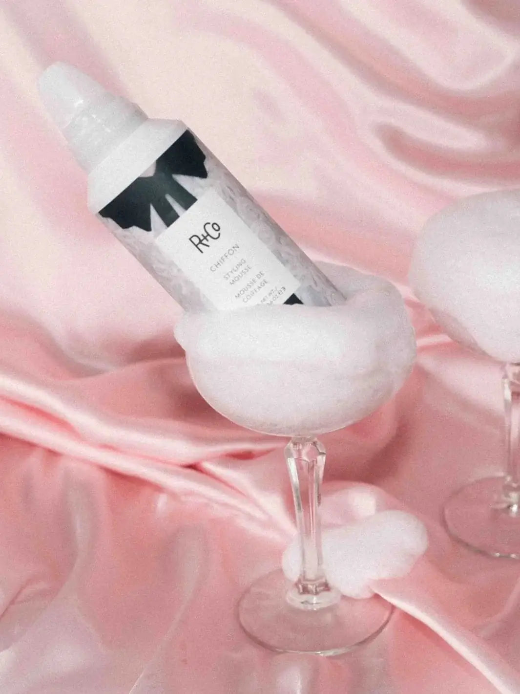 A bottle of R+Co CHIFFON Styling Mousse rests inside a coupe glass filled with fluffy white foam against a pink satin background | Twentyseven Toronto