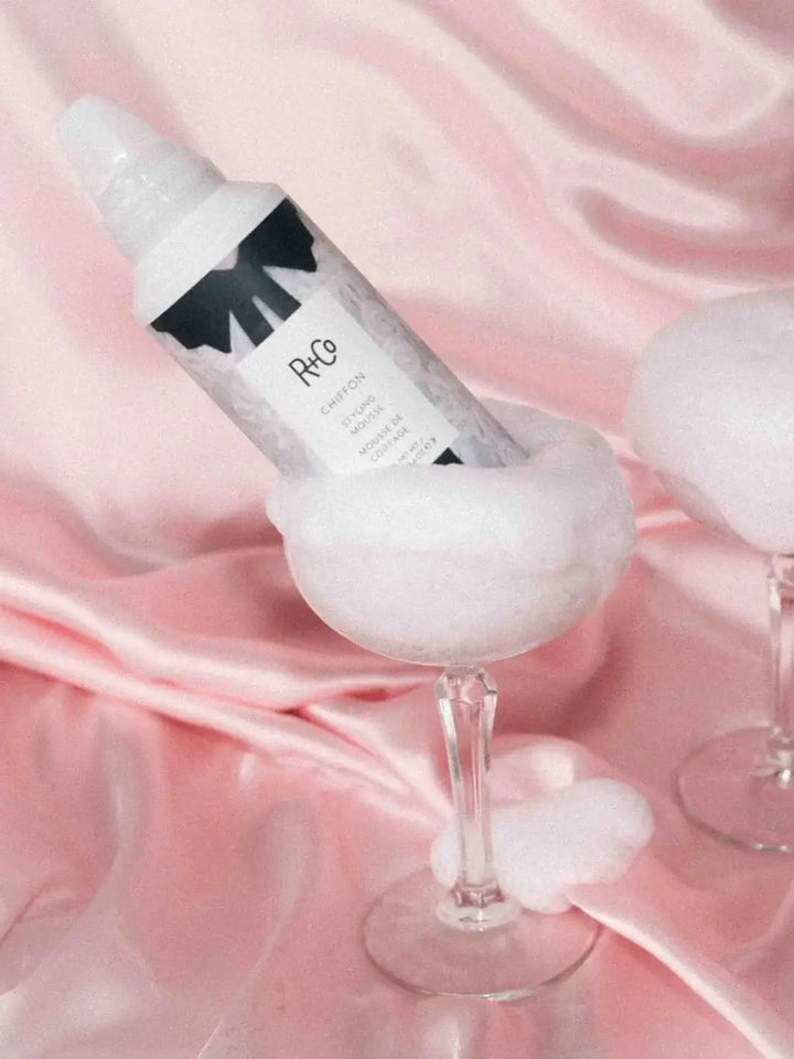 A bottle of R+Co CHIFFON Styling Mousse rests inside a coupe glass filled with fluffy white foam against a pink satin background | Twentyseven Toronto