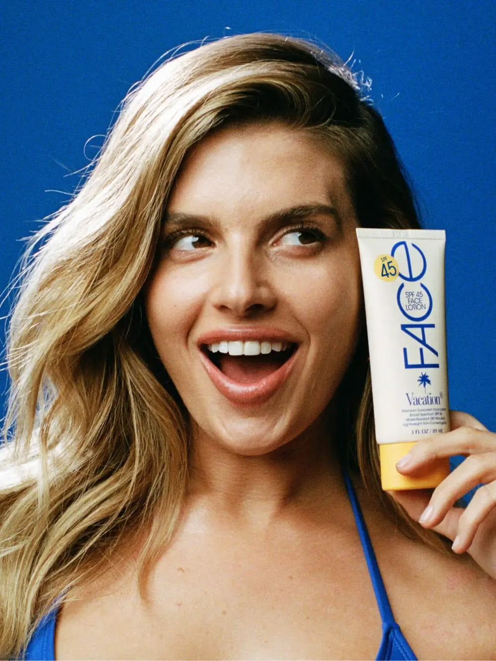 A blonde woman smiling while holding a tube of Vacation Inc Classic Face Lotion SPF 45 up to her face against a blue background | Twentysevn Toronto