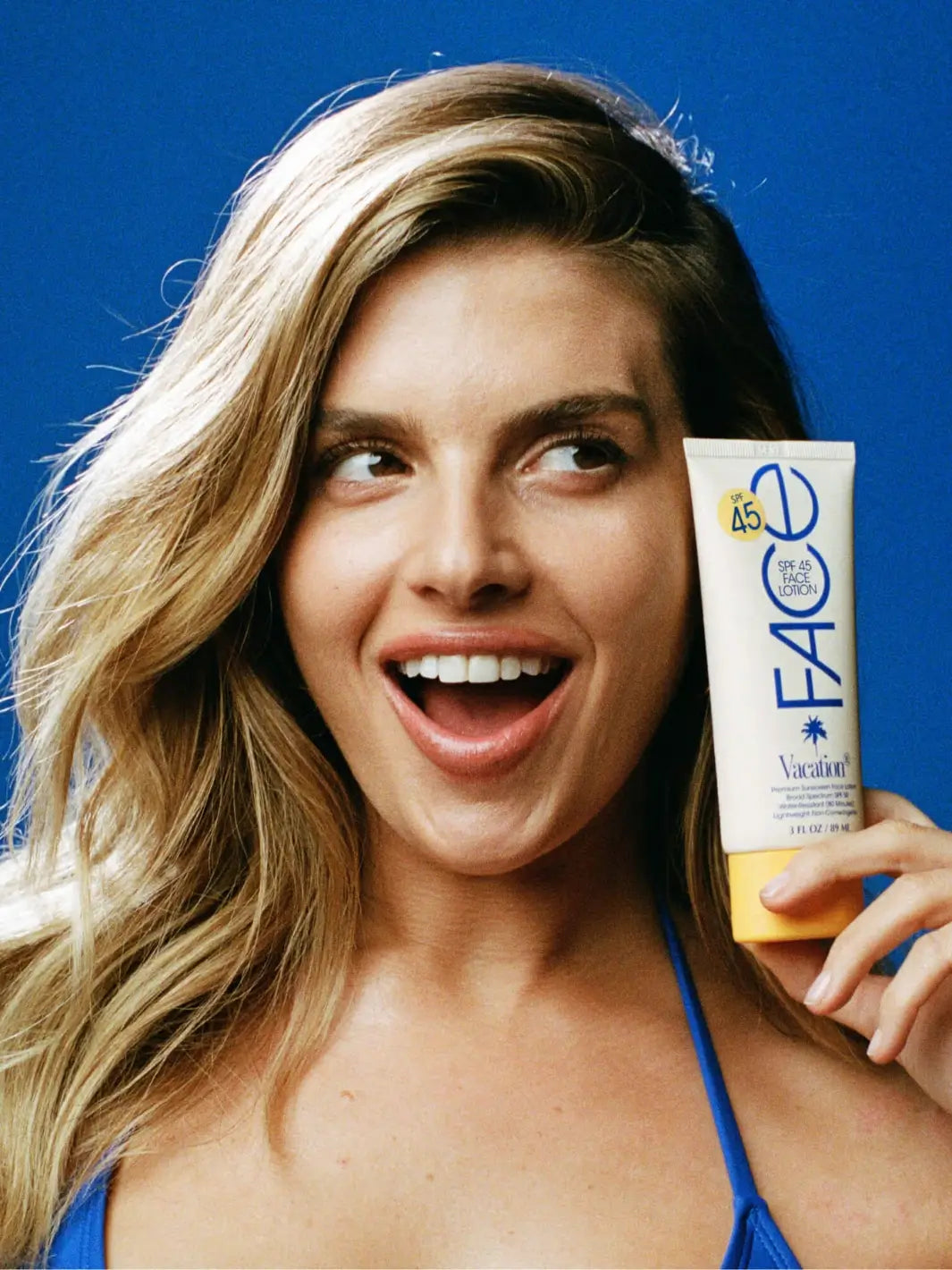 A blonde woman smiling while holding a tube of Vacation Inc Classic Face Lotion SPF 45 up to her face against a blue background | Twentysevn Toronto