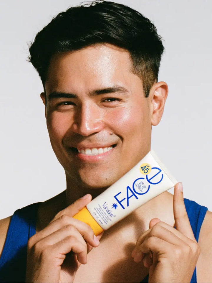 A man smiling while holding a tube of Vacation Inc Classic Face Lotion SPF 45 horizontally below his chin | Twentyseven Toronto