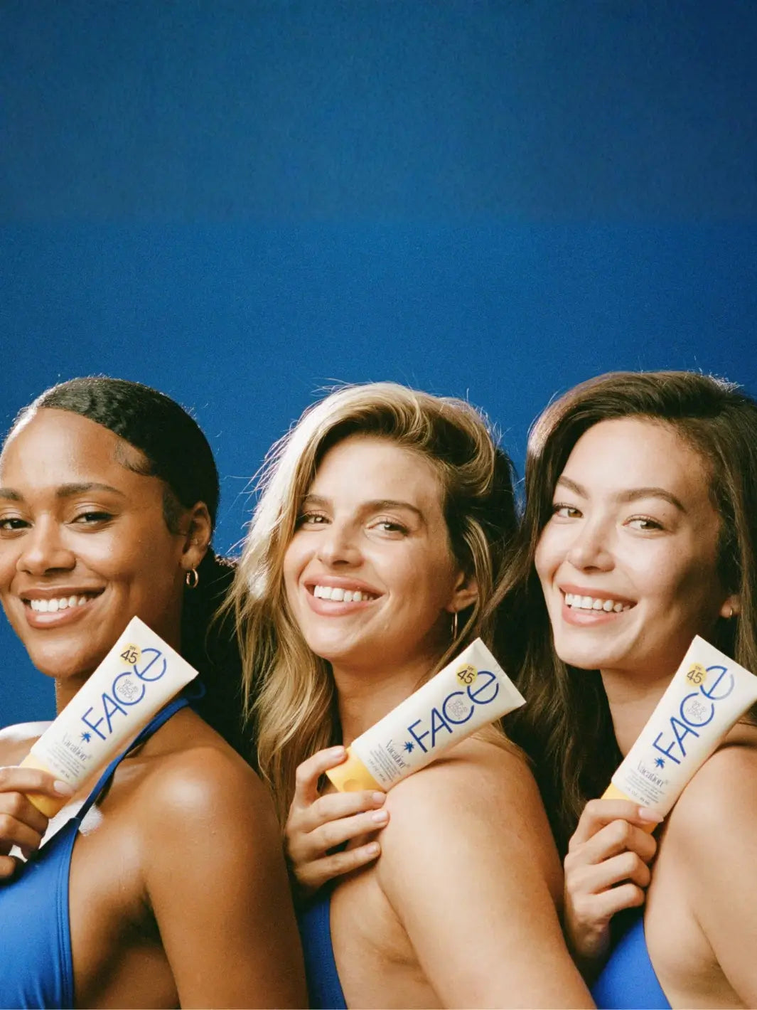 Three smiling women with diverse skin tones stand shoulder-to-shoulder, each resting a tube of Vacation Inc Classic Face Lotion SPF 45 against their skin | Twentyseven Toronto