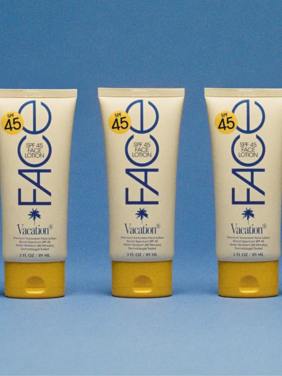 Three identical tubes of Vacation Inc Classic Face Lotion SPF 45 stand upright in a neat row against a textured blue background | Twentyseven Toronto