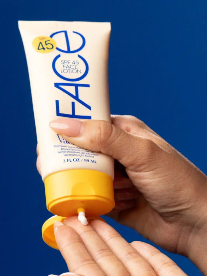 A hand squeezing a dollop of white cream from a tube of Vacation Inc Classic Face Lotion SPF 45 against a blue background | Twentyseven Toronto