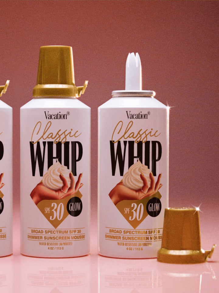 Vacation Inc. Classic Whip Glow SPF 30 | Twentyseven Toronto | Two cans of 'Vacation Classic Whip' sunscreen with gold nozzles on a pink background.