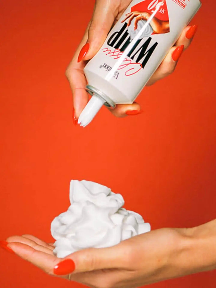 A close-up shot shows hands with bright red nail polish dispensing a dollop of white sunscreen mousse from a Vacation Inc Classic Whip SPF 50 can against a vibrant red background | Twentyseven Toronto