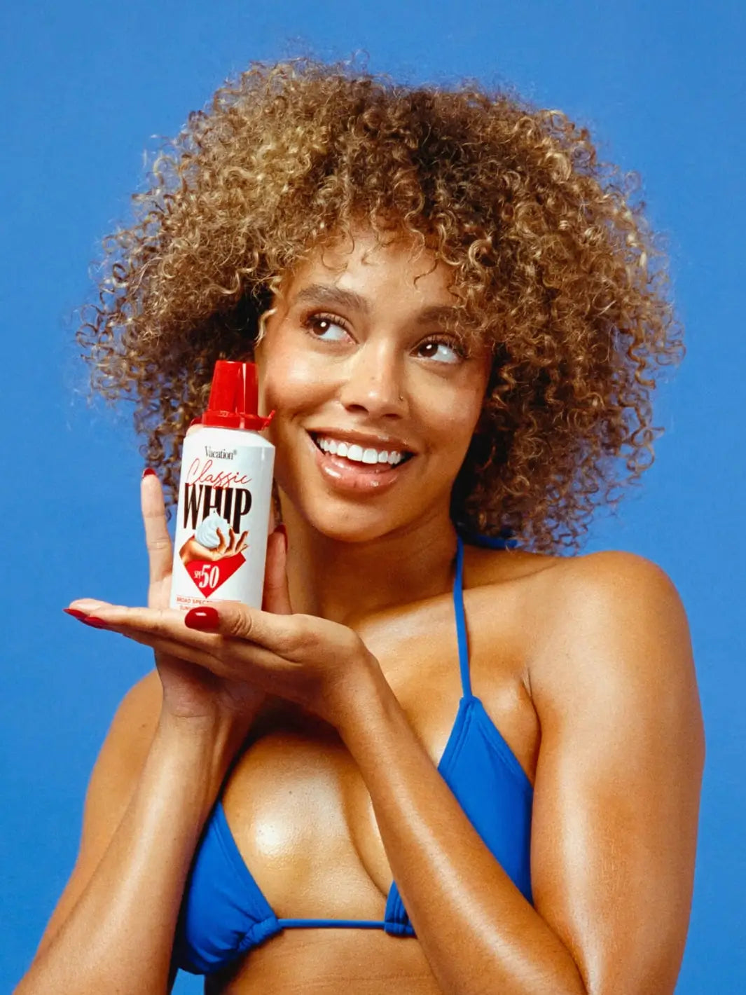 A woman with curly hair poses in a blue bikini while presenting a bottle of Vacation Inc Classic Whip SPF 50 on her open palm against a bright blue background | Twentyseven Toronto