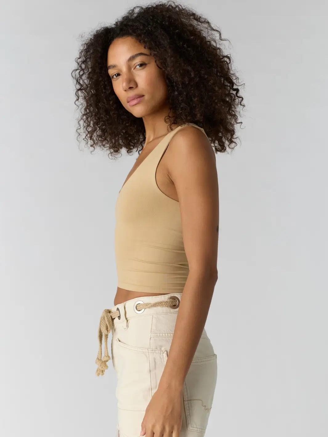 Woman with curly hair wearing a beige 'Clean Lines Muscle Cami' fitted top in 'Oat Latte' from Free People, side view | Twentyseven Toronto