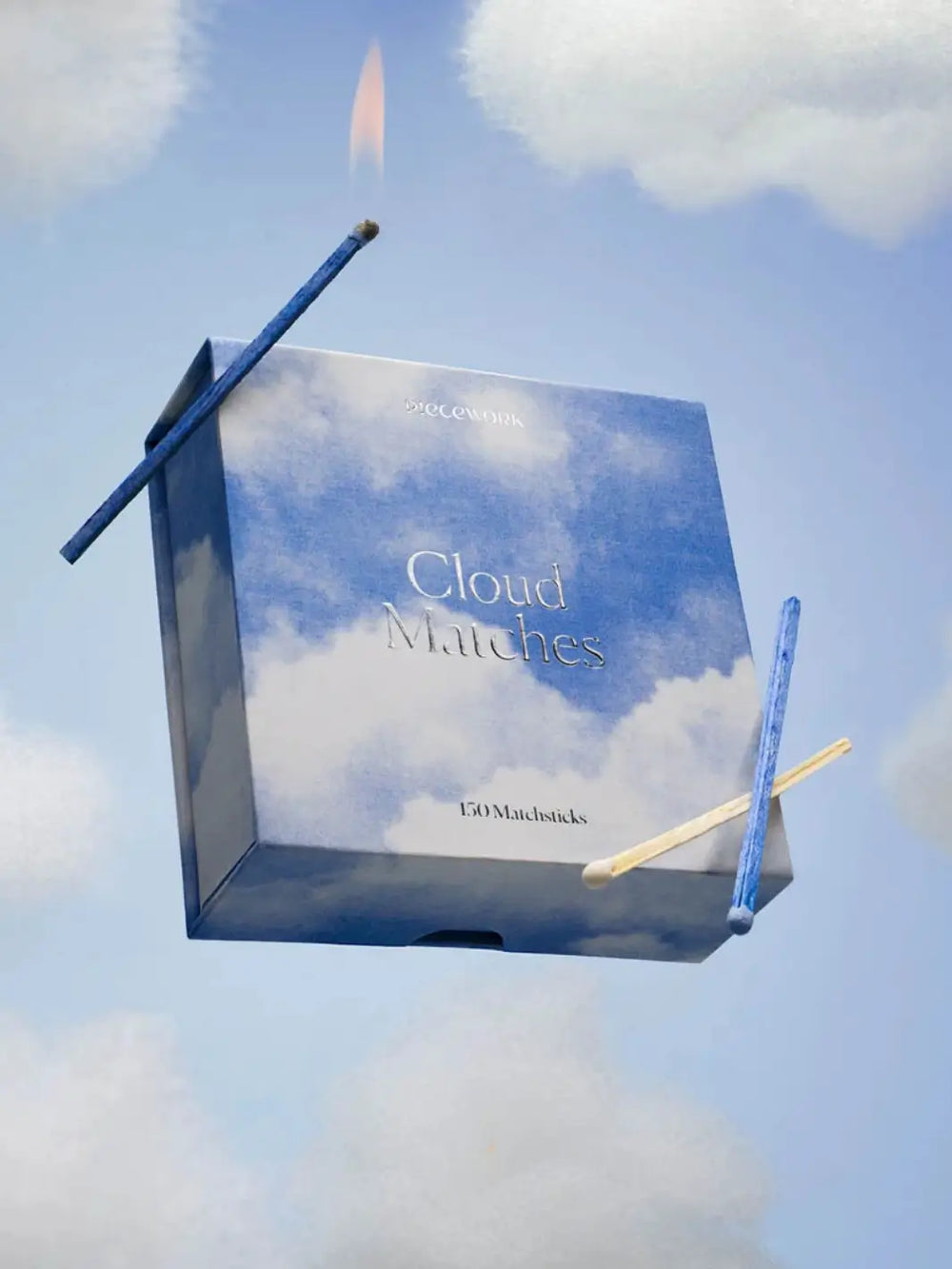 Box of matches with blue cloud design labeled 'Cloud Matches' from Piecework Puzzles with two lit matchsticks in the corner on a blue sky background | Twentyseven Toronto