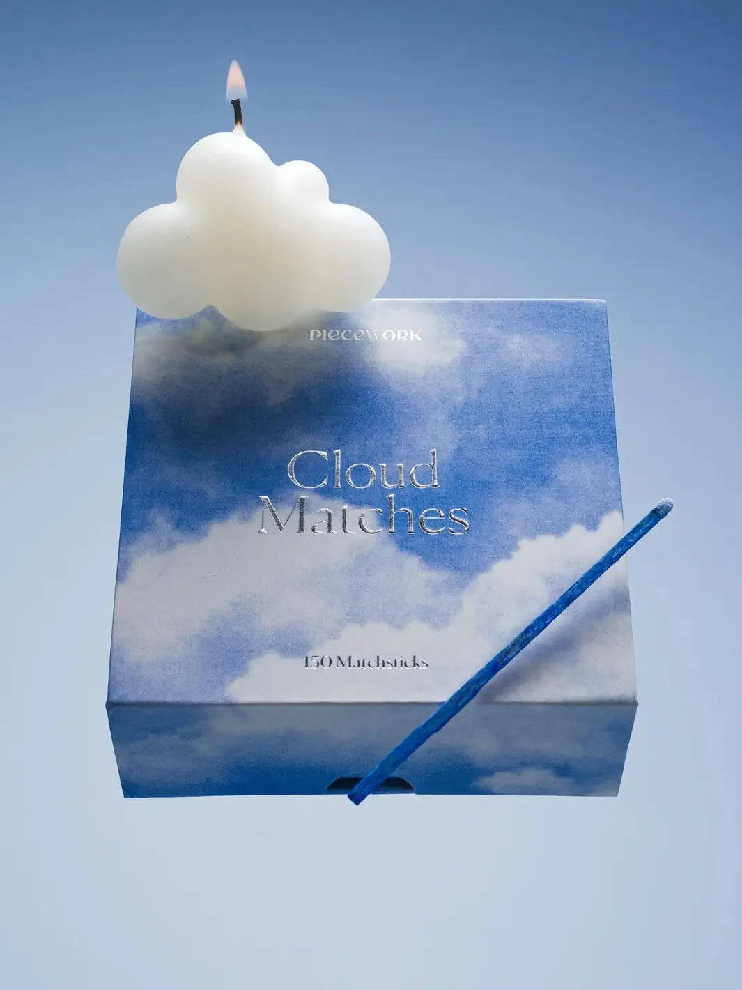 Box of matches with blue cloud design labeled 'Cloud Matches' from Piecework Puzzles with a lit cloud shaped candle on a light blue background | Twentyseven Toronto