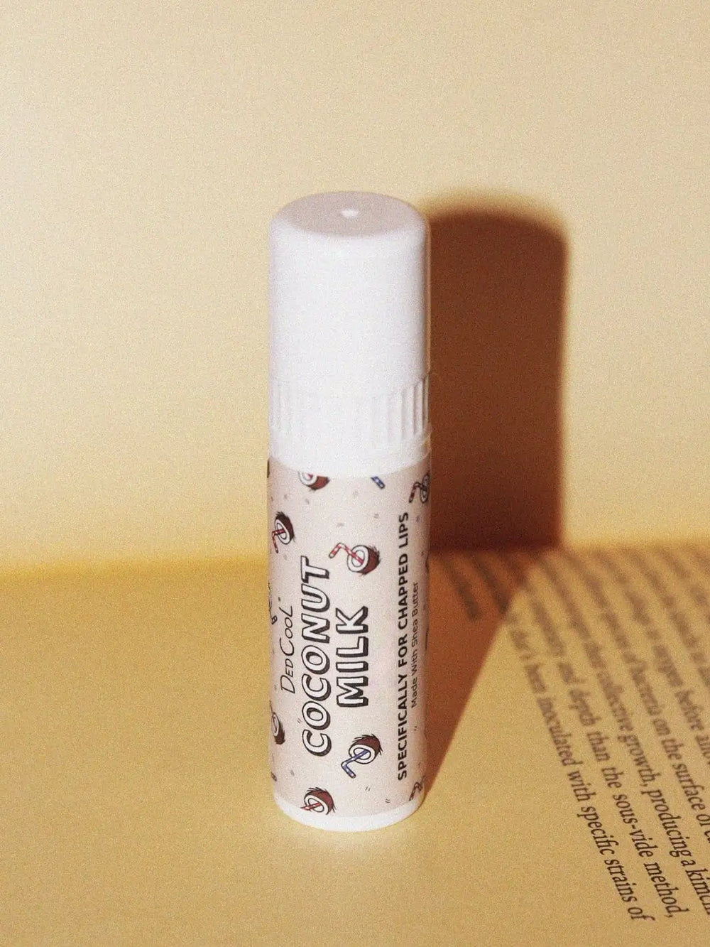 A tube of Dedcool Coconut Milk Balm Stick stands on an open book, casting a shadow against a warm cream background | Twentyseven Toronto
