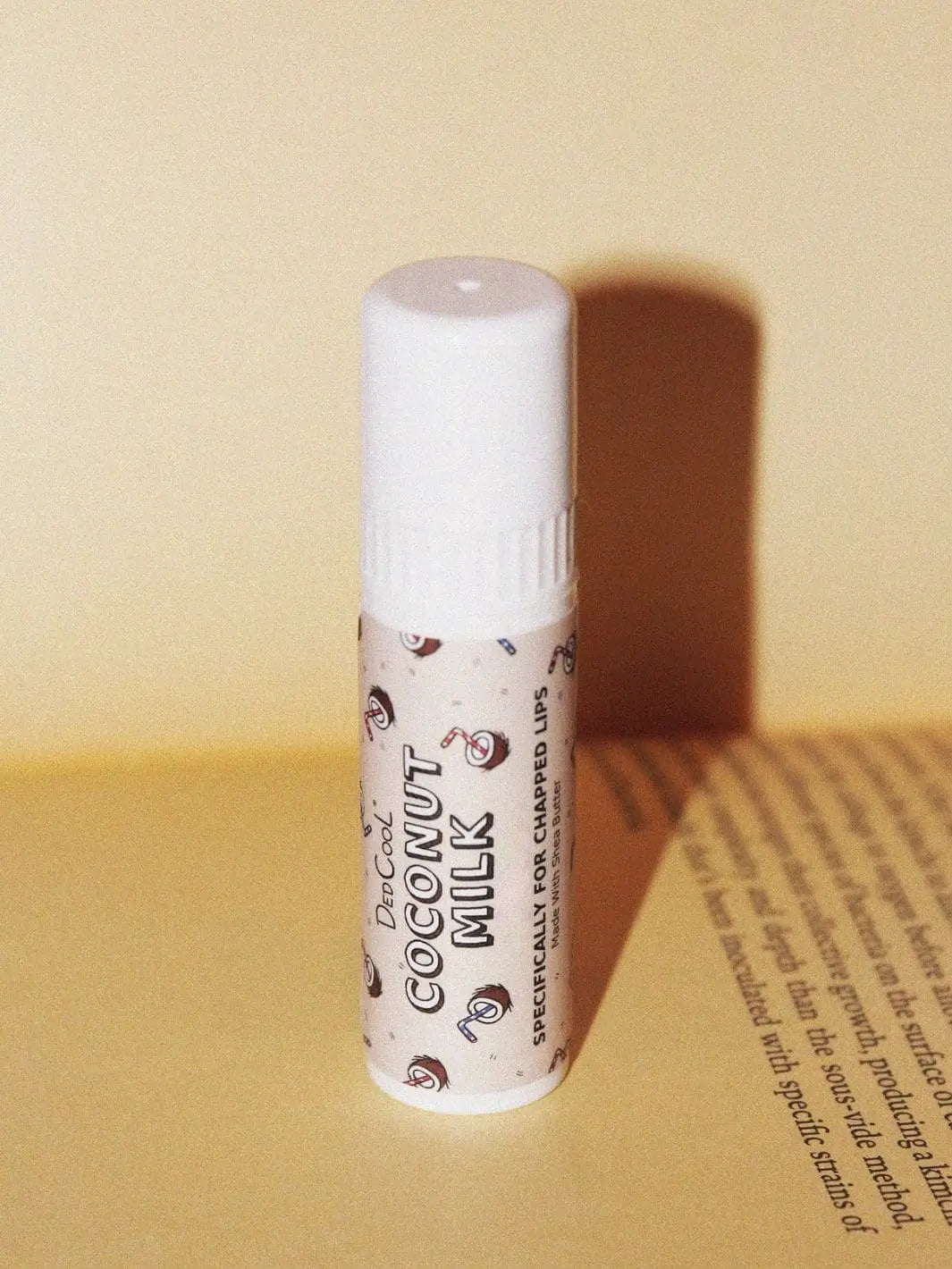 A tube of Dedcool Coconut Milk Balm Stick stands on an open book, casting a shadow against a warm cream background | Twentyseven Toronto