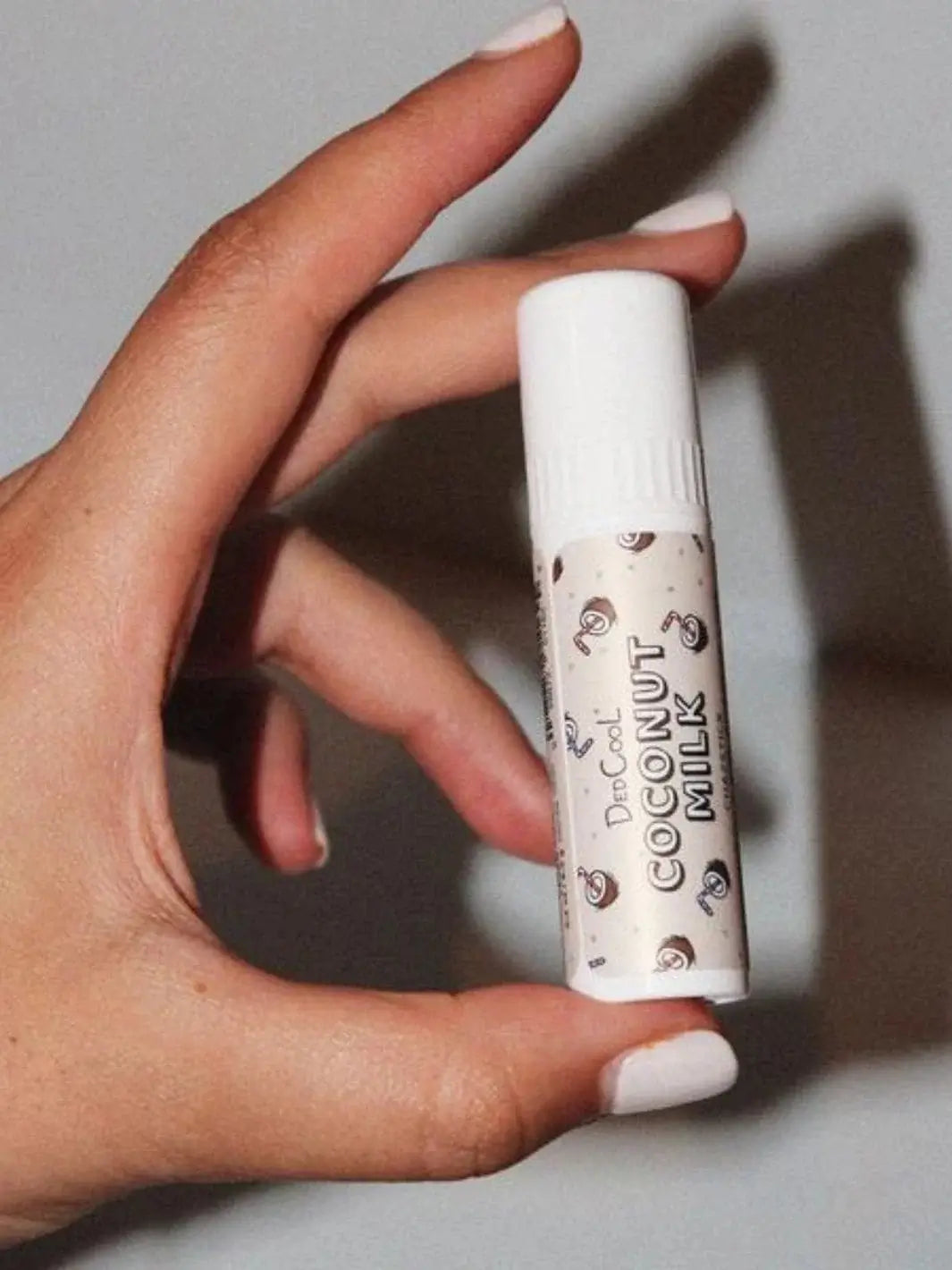 A hand with light-colored fingernails holds a tube of Dedcool Coconut Milk Balm Stick vertically against a plain gray background | Twentyseven Toronto