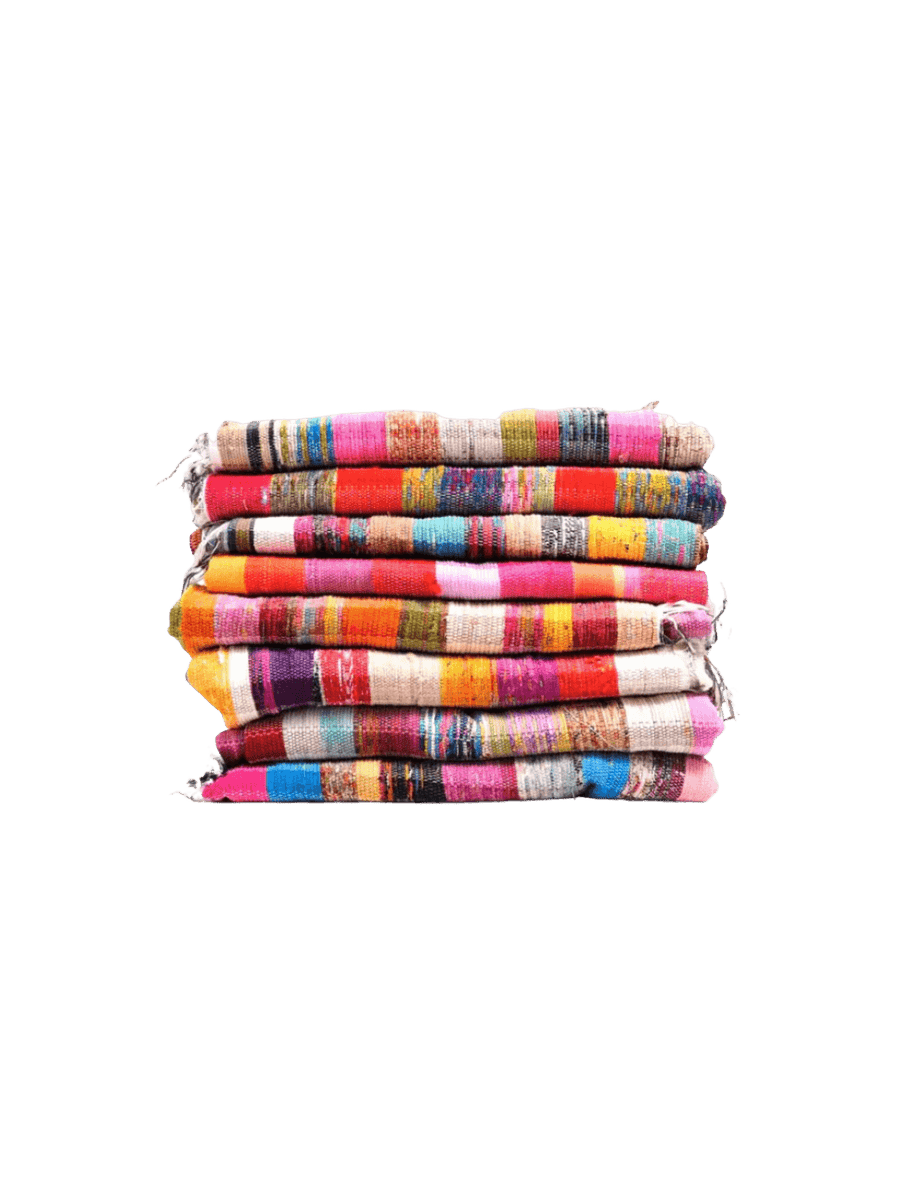 A tall stack of eight Collected Rug from Puebco textiles displays a vibrant array of multi-colored bohemian stripes, rich textures, and rustic, frayed edges | Twentyseven Toronto