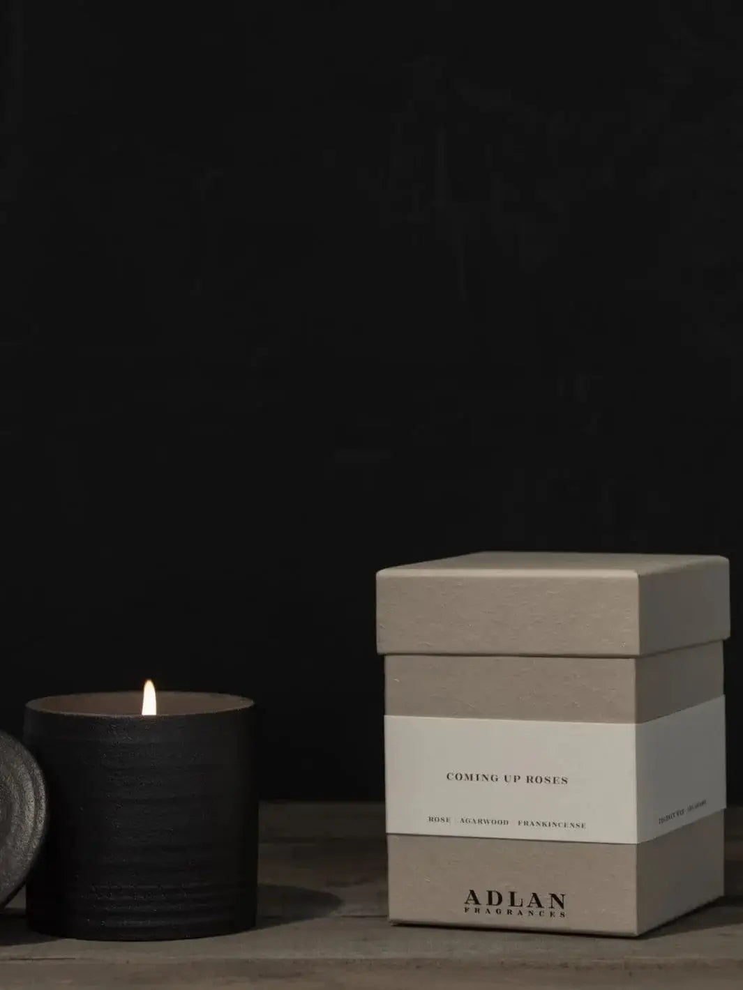 A lit Adlan Fragrances Coming Up Roses - Petite candle glows softly inside its ribbed black jar next to its grey paper packaging box | Twentyseven Toronto