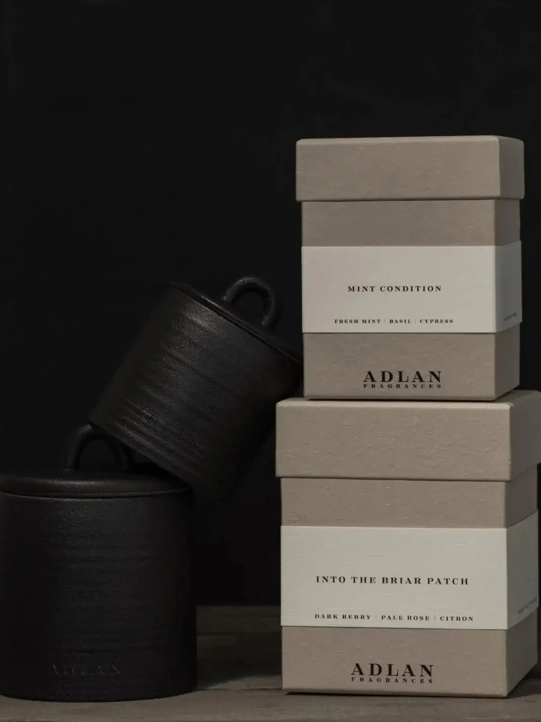 Two black lidded jars, one small, one large, are stacked alongside two grey product boxes for a dynamic display of alternative options to the Adlan Fragrances Coming Up Roses - Petite candle line | Twentyseven Toronto