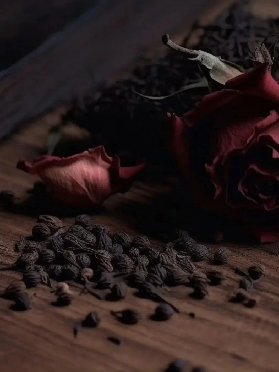 A moody, atmospheric close-up features a dried red rose, a single petal, and a scattering of natural botanicals on a wooden surface, representing the scent inspiration for the Adlan Fragrances Coming Up Roses - Petite candle | Twentyseven Toronto