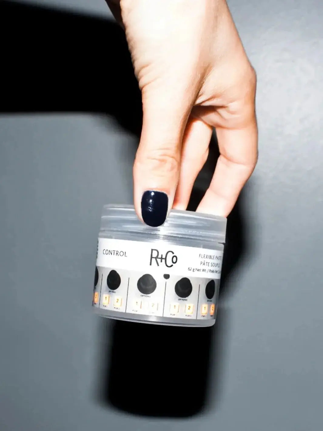A hand with dark nail polish holds a small clear jar of R+Co CONTROL Flexible Control Paste against a gray background | Twentyseven Toronto