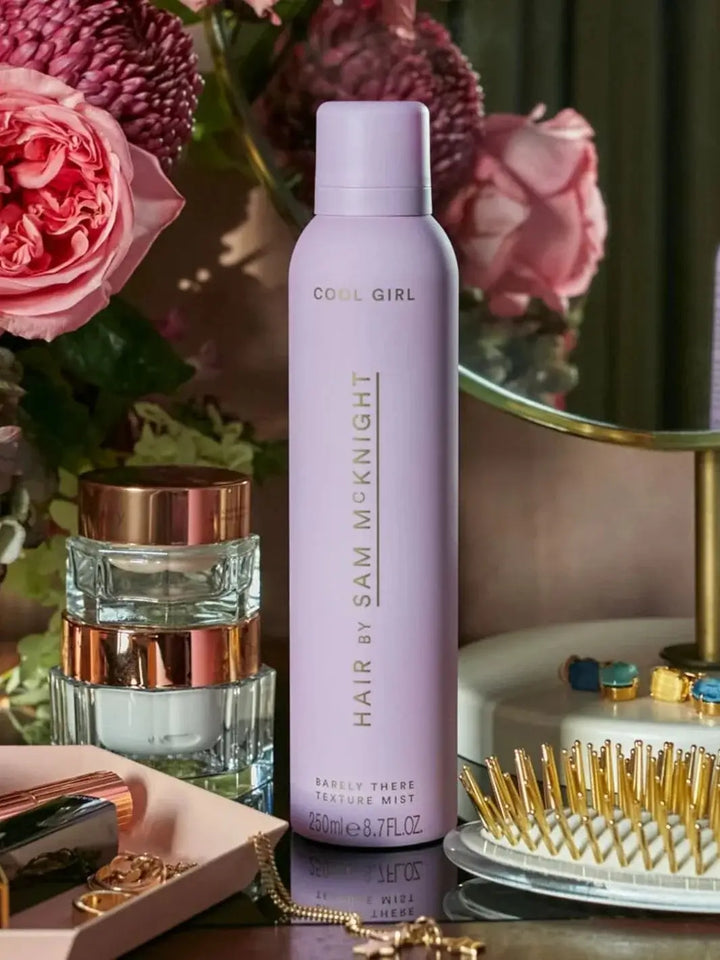 A pastel pink aerosol can sits on a dark vanity table next to luxury skincare jars and a gold-bristled hairbrush to elevate Hair by Sam McKnight Cool Girl Barely There Texture Spray | Twentyseven Toronto