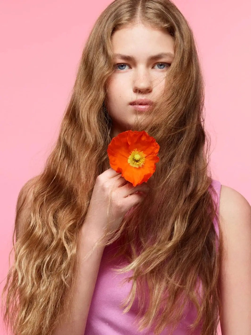 A young woman with textured, piecey blonde waves holds a bright orange poppy flower against a solid pink background to reflect the fresh aesthetic of Hair by Sam McKnight Cool Girl Barely There Texture Spray | Twentyseven Toronto