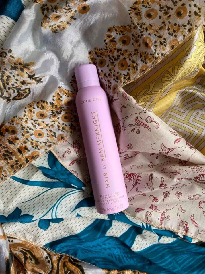 A purple spray bottle is styled flat-lay on top of an array of patterned silk scarves to accentuate the artistic design of Hair by Sam McKnight Cool Girl Barely There Texture Spray | Twentyseven Toronto