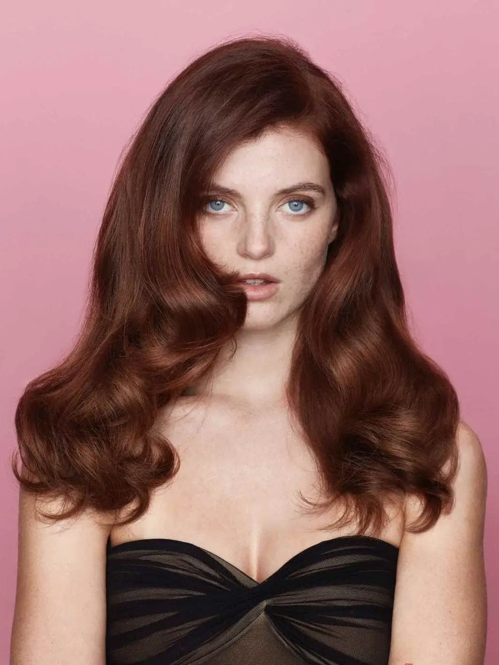 A woman with auburn hair displays a thick, wavy blowout style with lifted roots against a pink backdrop, demonstrating the styling power of Hair by Sam McKnight Cool Girl Volume Foam | Twentyseven Toronto