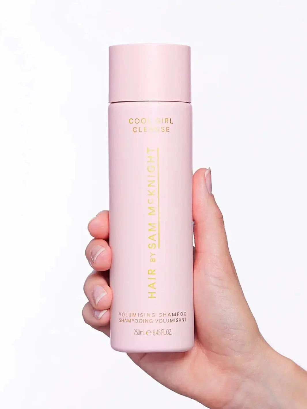 A hand holds up a tall, pastel pink bottle of Hair by Sam McKnight Cool Girl Volume Shampoo against a plain white background | Twentyseven Toronto