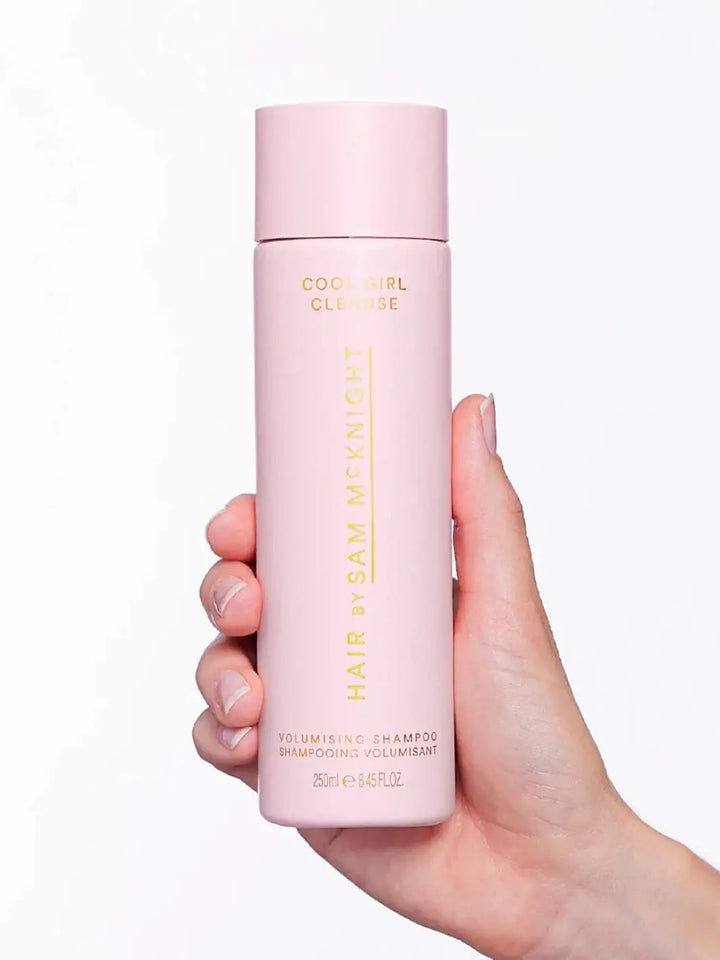 A hand holds up a tall, pastel pink bottle of Hair by Sam McKnight Cool Girl Volume Shampoo against a plain white background | Twentyseven Toronto
