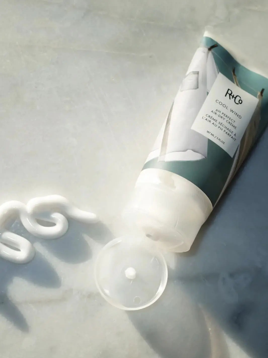 Tube of R+Co 'COOL WIND pH Perfect Air Dry Creme' next to a product swatch on a a marble background, 147ml | Twentyseven Toronto